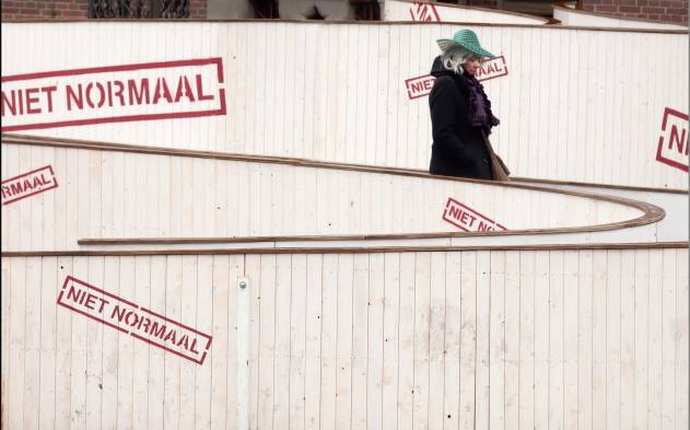 Vrouw met groene hoed loopt op kronkelende hellingbaan waarop woorden Niet Normaal staan.